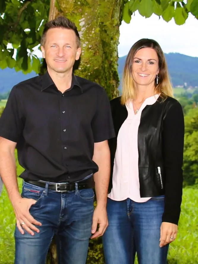 PIT Geschäftsführung stehen lächelnd im Grünen vor einem Baum, halbnahes Porträtfoto im Querformat.